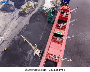 Workers are loading coal onto a cargo ship at a busy port. Cranes and machinery are in use to transfer the coal from the ground to the ship. It is daytime and many activities are happening.