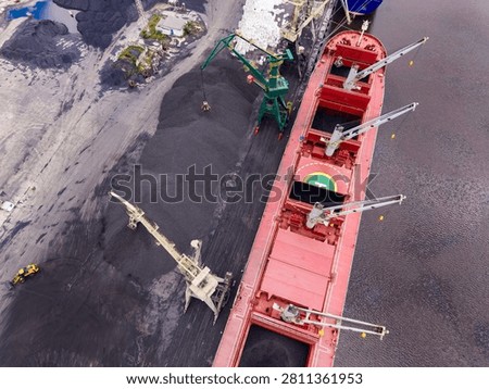Workers are loading coal onto a cargo ship at a busy port. Cranes and machinery are in use to transfer the coal from the ground to the ship. It is daytime and many activities are happening.