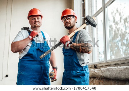 Workers, looking at the camera, standing at the construction site. They are wearing red safety helmets and one of them is holding a sledgehammer for repairs.