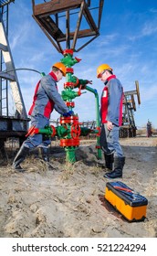 Workers In The Oilfield, One Repairing Wellhead With The Wrench, Other Supervising. Pump Jack And Wellhead Background. Oil And Gas Concept. 