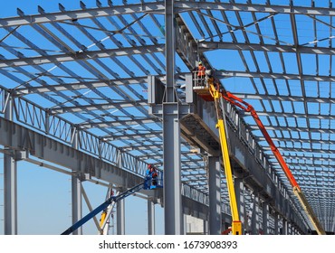 Workers on aerial work platforms build the metal structure of the roof of a large building.