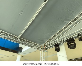 Workers prepare a stage area by setting up a large canopy and arranging lighting equipment. The event is set indoors with tall ceilings and a spacious layout.