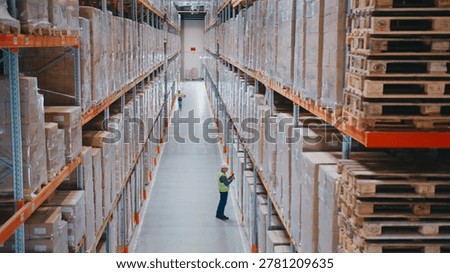 Workers in protective helmets moving between massive storage shelves filled with boxes. Two employees carrying package. Employees walking or inspecting stacked inventory along warehouse aisle.