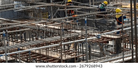 Workers in safety gear are constructing a large building. They are using steel rods and scaffolding as they work high above the ground at a bustling construction site