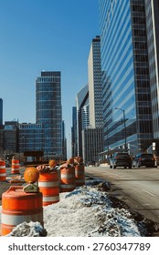 Workers set up construction barriers during the day on a street filled with tall buildings. Vehicles pass by as the skyline is visible above the work area.