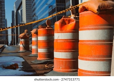 Workers set up construction barriers on a city street lined with tall buildings. Orange barrels and caution tape keep pedestrians away from the work area. It is daytime, and the street is busy.