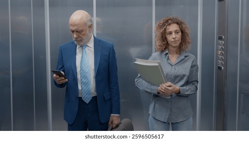 Workers standing in the elevator, a man is checking his smartphone and the woman is carrying paperwork