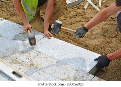 Workers Tapping Pavers Into Place With Rubber Mallets. Installation Of Granite Paver Blocks Series With Motion Blur On Hammers And Hands.