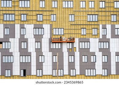 Workers team on construction cradle cladding and warming wall of monolithic house under construction. Man on suspended platform insulating wall facade with rock wool and installing ventilated facade - Shutterstock ID 2345369865