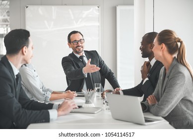 Workers And Their Chief Sitting At The Table. Chairman Showing With Hand At Attendee And Smiling