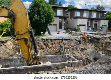 Workers In Trench Looking At Excavator Digging Earth Out Of A Trench Box For Laying New Sewer Lines On Residential Street Nepean, Ontario, Canada - July 23, 2020