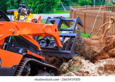 Workers are using compact track loader to extract large tree stump from backyard in suburban neighborhood.