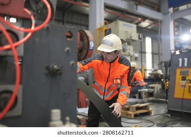 Workers are using large machines in a factory. They wear helmets and bright clothing. The scene is busy with machines running and people engaged in their work.
