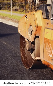 Workers are using a large steamroller to compact fresh asphalt on a newly paved road. The sun is shining, highlighting the vibrant surroundings and the meticulous work being done outdoors.