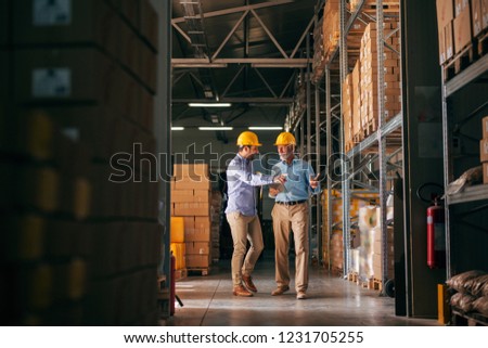 Workers walking in storage and pointing at boxes. In hands folder and tablet, on heads helmets.