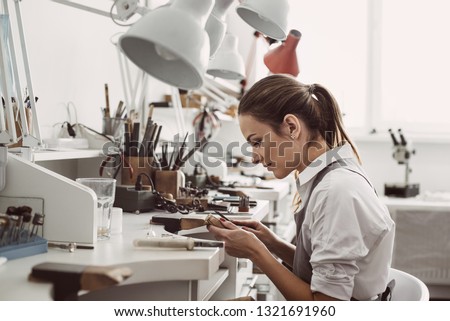 Working all day. Side view of young female jeweler sitting at her jewelry workshop and holding in hands jewelry tools for making accessories