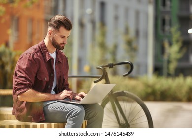 Working anywhere he wants. Side view of young man with stubble in casual clothes working on laptop while sitting on the bench near his bicycle