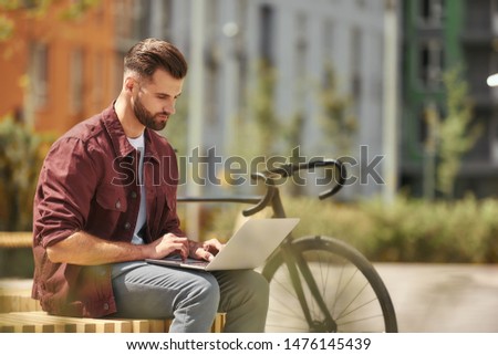 Working anywhere he wants. Side view of young man with stubble in casual clothes working on laptop while sitting on the bench near his bicycle