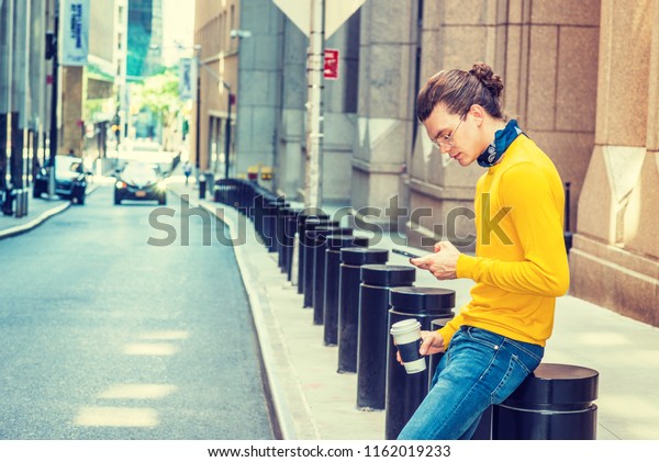 Working Break. Hispanic American Man with hair\
bun, wearing glasses, yellow long sleeve T shirt, jeans, small\
black scarf around neck, holding cup of coffee, sitting on street,\
texting on cell phone.
