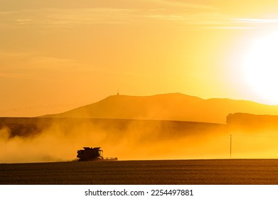 Working combine harvester to dump grain from its loader in a wheat field in the summer months during sunset. Clouds of dust behind. - Shutterstock ID 2254497881