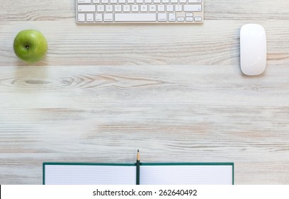 Working desk with green apple. View from above on light beige wooden working desk with green apple, large notepad, computer keyboard and mouse, green pencil