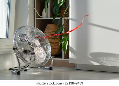 Working Electric Fan With Red Wriggle Ribbon Designed To Cool Air And Supply Wind Located Near Window Next To House Plant. Equipment For Cooling Temperature In Room In Hot Summer Weather