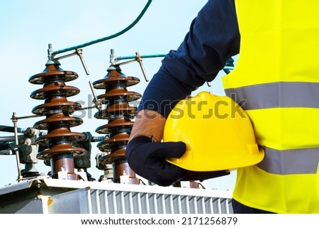 A working electrician in a yellow vest holds a protective helmet in his hand against the background of a high-voltage transformer and electrical substation equipment.