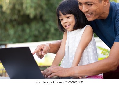 A Working Father And An Asian Kindergarten Cute Daughter, He Sits Teaching Her With A Smiling And Happy Face. Through A Black Notebook Computer Make Her Homeschooling Fun And Have Good Connections.