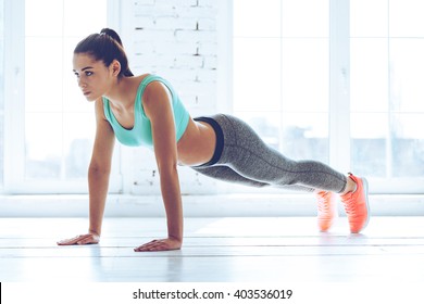 Working her core muscles. Full length of young beautiful woman in sportswear doing plank while standing in front of window at gym