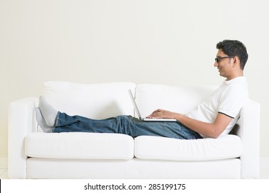 Working From Home Concept. Indian Guy Using Laptop Computer. Asian Man Relaxed And Sitting On Sofa Indoor. Handsome Male Model.