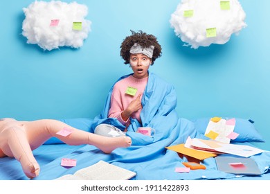 Working At Home During Quarantine. Surprised Curly African American Woman In Pajama Surrounded With Many Papers Poses In Bed With Inflated Doll Near Has Imaginary Friend During Self Isolation