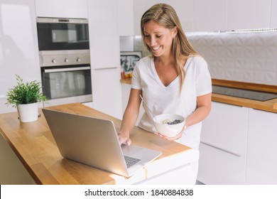 Working At Home. Happy Young Caucasian Woman In White T-shirt  Sitting At The Table With Laptop Computer. Kitchen At The Background.