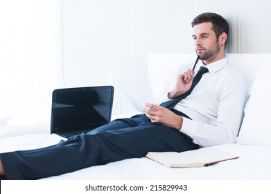 Working In Hotel Room. Thoughtful Young Man In Shirt And Tie Examining Document While Lying In Bed At The Hotel Room 