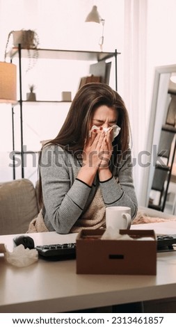 Working ill. Seasonal cold. Contagious disease. Office sickness. Young woman in blanket suffering from runny nose with tissue at face at table with keyboard and mug.