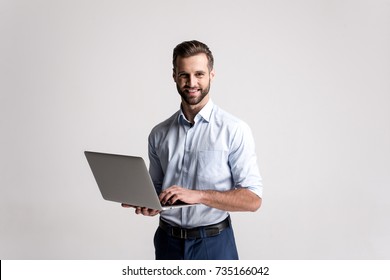 Working With Joy. Handsome Young Man Using His Laptop And Looking At Camera With Smile While Standing Against White Background.