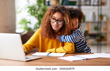 Working with kids. Young focused woman mother wearing eyeglasses using laptop and thinking about work task while small boy son gently hugs her. Childcare concept
