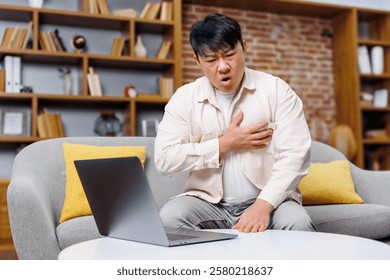 working man, having heart attack, chest heartache, Concerned man sitting on a couch experiencing chest discomfort while looking at a laptop in a modern living room