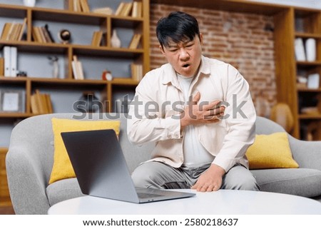 working man, having heart attack, chest heartache, Concerned man sitting on a couch experiencing chest discomfort while looking at a laptop in a modern living room