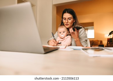 Working Mom Making Notes During A Phone Call With Her Clients. Multitasking Mom Working On A New Creative Project In Her Home Office. Female Interior Designer Carrying Her Baby On Her Laps.