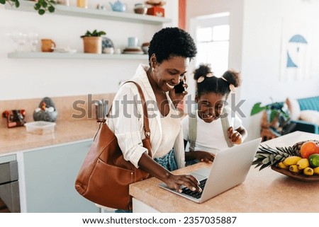 Working mom smiles while making a phone call and using her laptop in the morning, with her daughter standing by. Happy black woman balancing between work and parenthood in the digital age.
