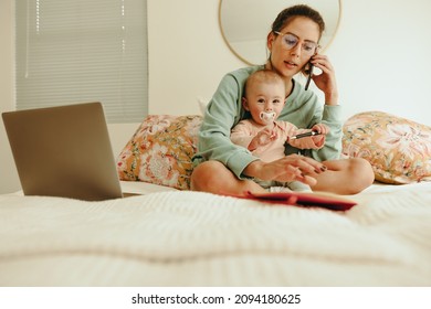 Working Mom Taking A Phone Call While Sitting On A Bed With Her Baby. Single Mother Communicating With Her Clients While Working From Home. New Mom Balancing Work And Motherhood.