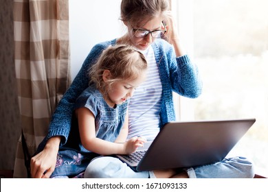 Working mom works from home office with kid. Mother and daughter read news. Woman and cute child sitting on window sill. Freelancer workplace with laptop and the Internet. Female business, career.