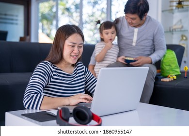 Working Mother Work From Home While Babysitting Her Playful Daughter At Home. Working Mother And Father Playing With Daughter Beside.