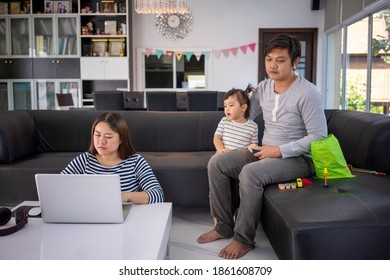 Working Mother Work From Home While Babysitting Her Playful Daughter At Home. Working Mother And Father Playing With Daughter Beside.