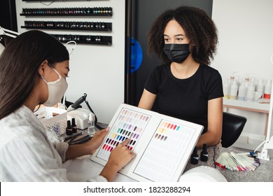 Working In Nail Salon During COVID-19 Epidemic. Asian Client Lady In Protective Mask Choose Color For Nails, African American Woman Looks At Clients In Interior Of Salon, Free Space