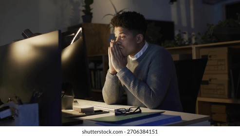 Working office worker wearing grey sweater at night office desk, with two monitors, lamp, notebooks. Workstation, screens, latenight, focus, calm, warmlight, shelves