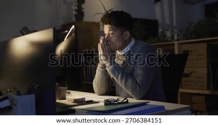 Working office worker wearing grey sweater at night office desk, with two monitors, lamp, notebooks. Workstation, screens, latenight, focus, calm, warmlight, shelves