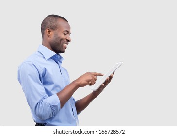 Working on digital tablet. Side view of handsome African man working on digital tablet and smiling while standing isolated on grey