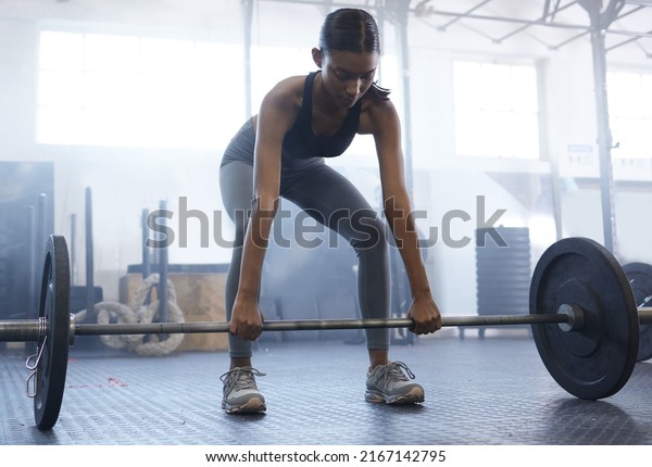 Working on her core\
strength and stability. Shot of a sporty young woman exercising\
with a barbell in a\
gym.