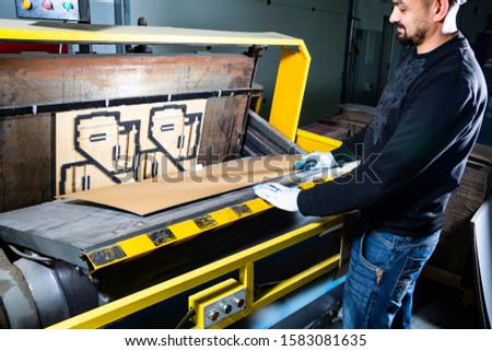 Working on a punching machine. Worker in a hard hat in cardboard boxes factory placing a carton in a paper die cutting machine.